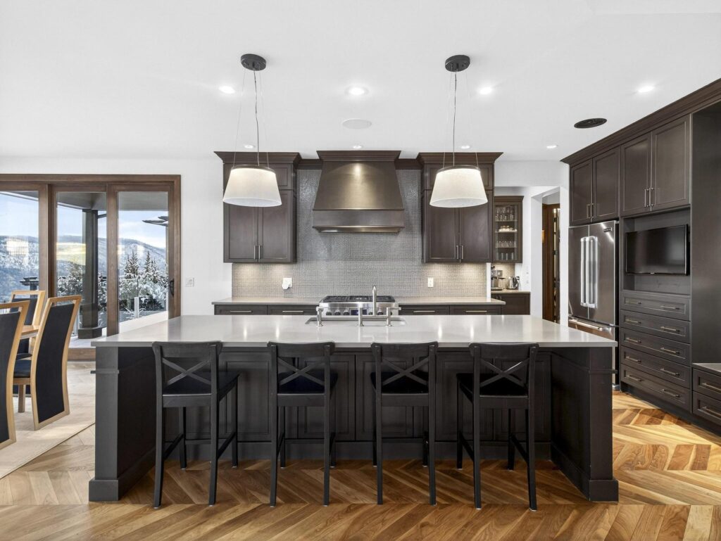 Kitchen with dark cabinetry, a range hood, pendant lighting and a long island with six stools, on herringbone wood floors.