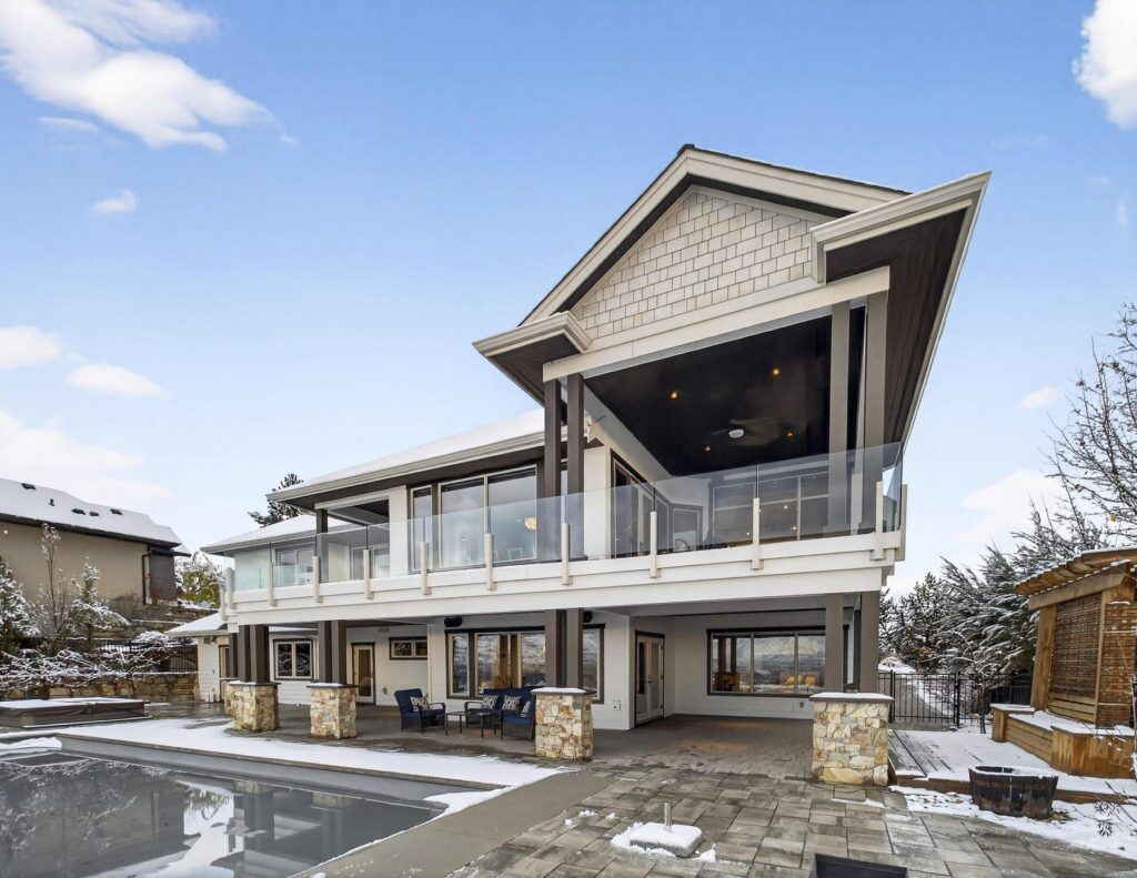 Rear of a large shingled home with a two-storey covered deck and glass railings, above a pool deck cleared of snow.