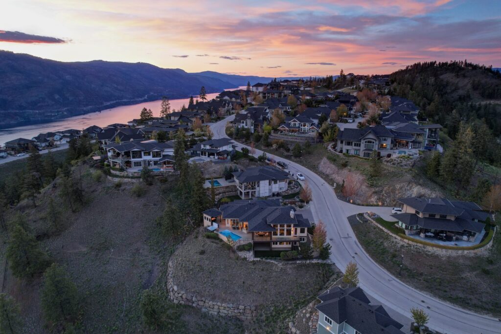 Aerial view at dusk over a hillside neighbourhood of large homes, with the lake and a pink sky beyond.