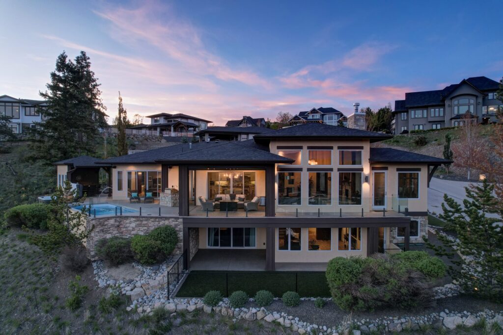 Rear of a contemporary hillside home at dusk, its windows lit, with a pink and blue sky over the ridge behind.