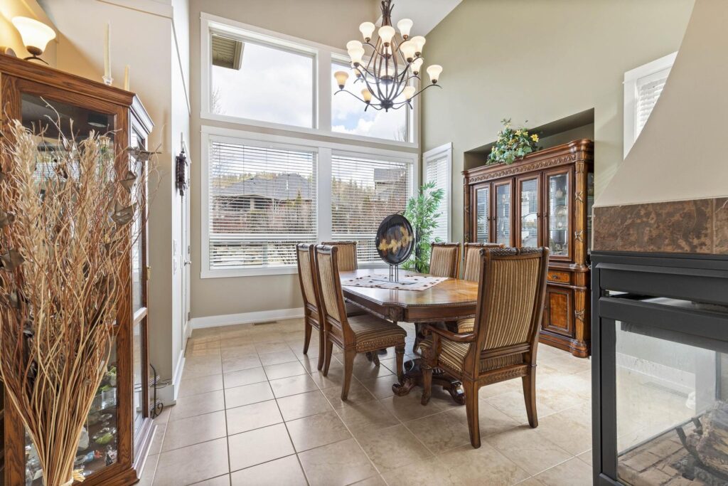 Dining area with a round table, a chandelier and tall windows, beside a tiled fireplace surround.