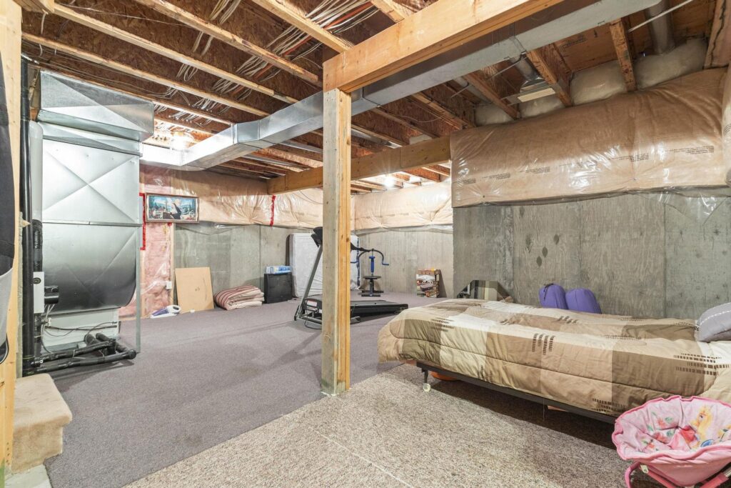 Unfinished basement with exposed floor joists overhead, bare framing, poly sheeting over the walls and belongings stored on the floor.