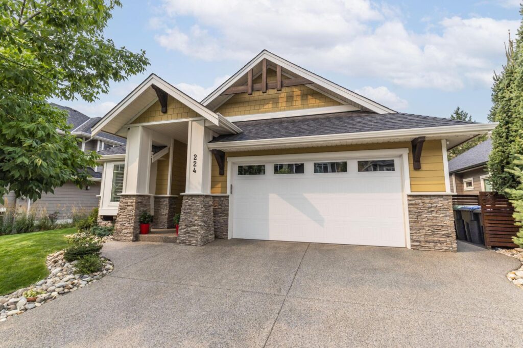 Front of a two-storey Craftsman-style home with a gabled roof, stone-clad columns and a double garage, with a concrete driveway and clipped shrubs.