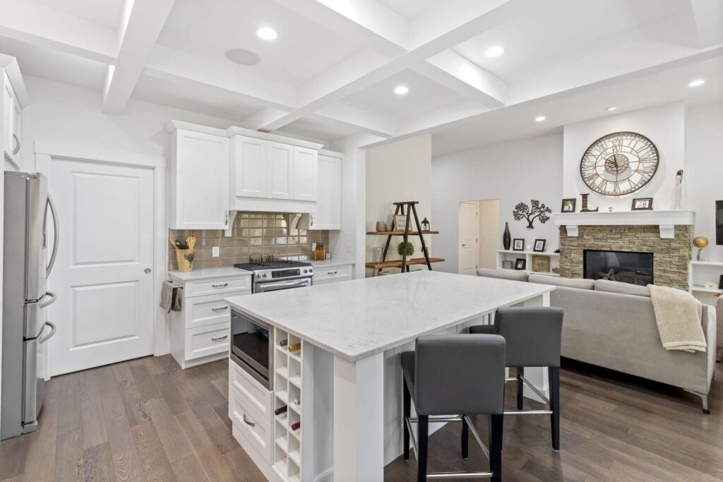 White kitchen with a large quartz island and two grey stools, a coffered ceiling, and a stone fireplace in the sitting area beyond.