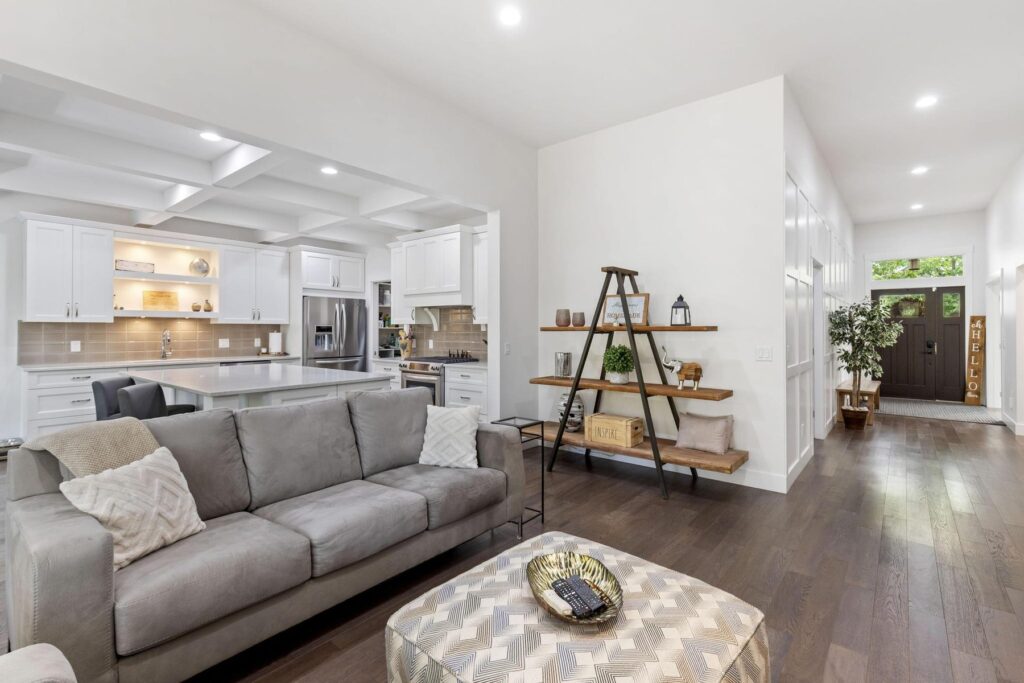 Open living area with a grey sectional sofa and patterned ottoman on dark hardwood, looking through to the kitchen and a door to the yard.