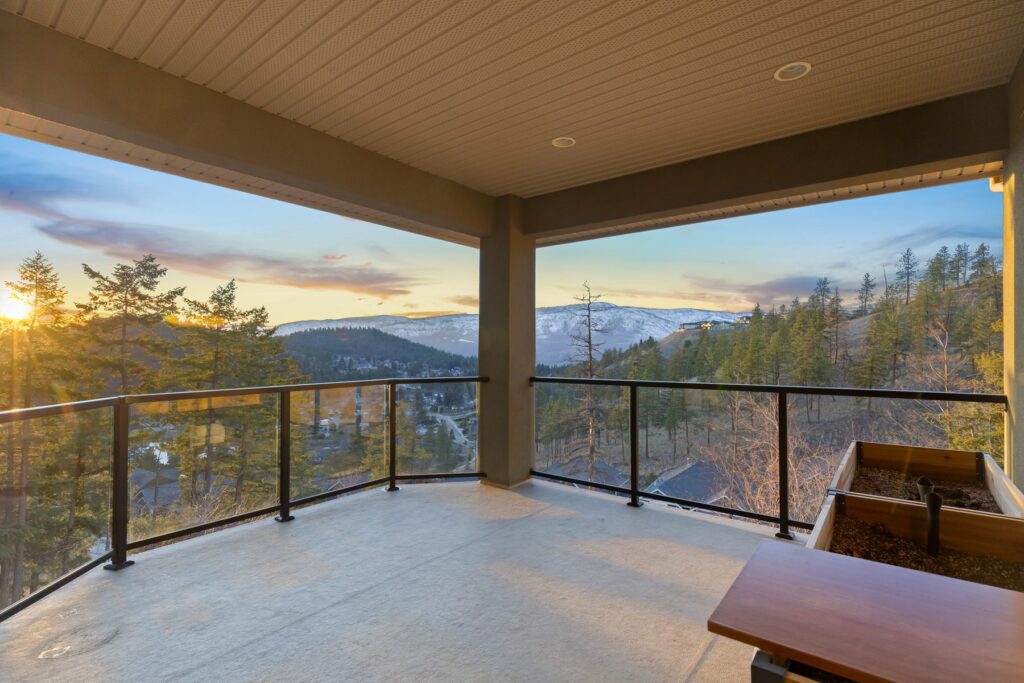 Covered deck with a wood ceiling and glass railing at sunset, looking across pines to hills and a low sun.
