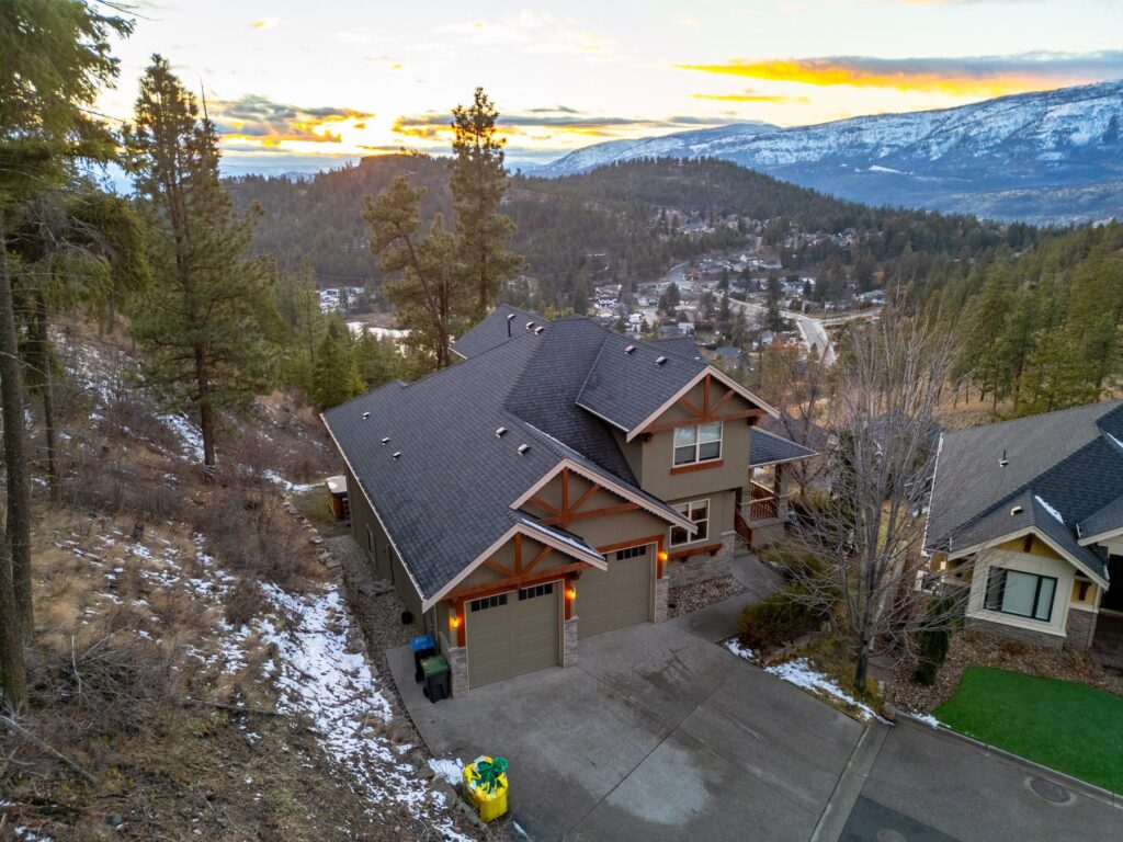 Aerial view at dusk of a house with lit windows on a treed slope, snow-capped mountains along the horizon.