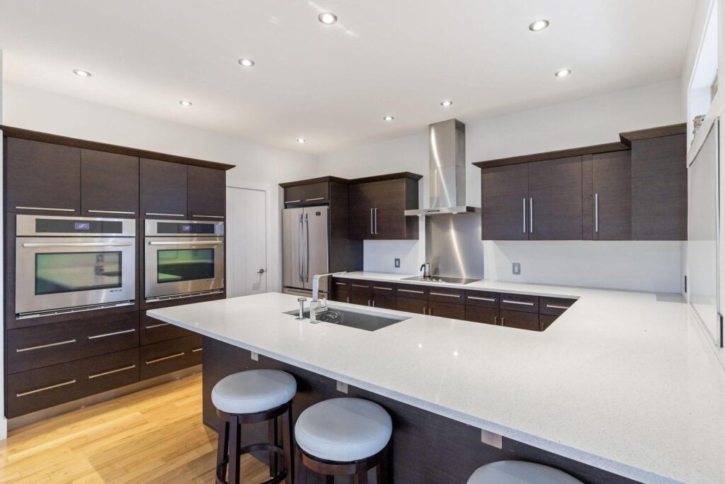 Kitchen with dark cabinetry, wall ovens, a stainless range hood and a long pale island with three stools.