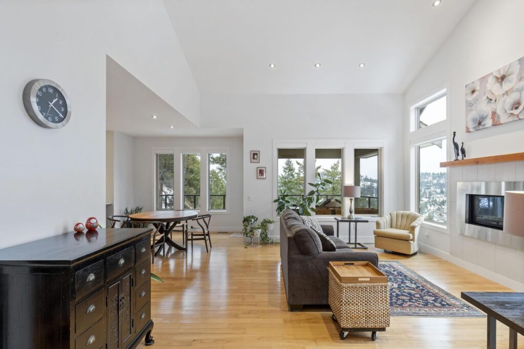 Vaulted open living and dining area with pale hardwood, a wall clock, and French doors to a deck.