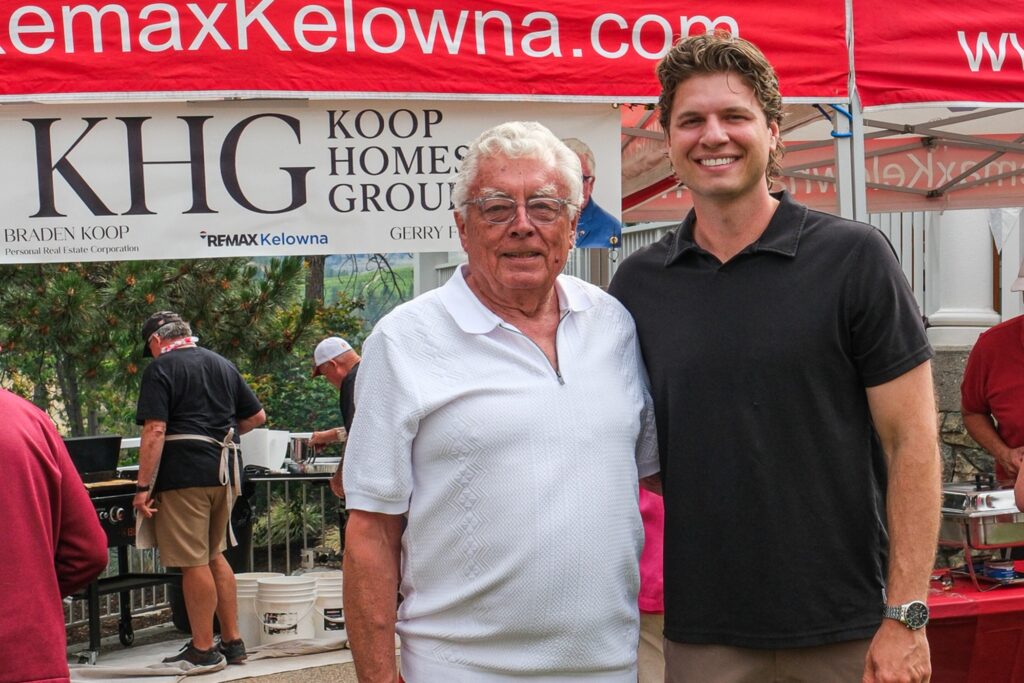 Braden Koop and Gerry Fraser at the RE/MAX Kelowna Canada Day pancake breakfast, in front of the Koop Homes Group banner