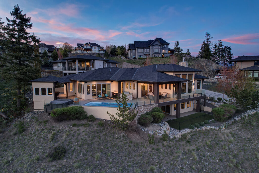 Twilight rear elevation of a Kelowna hillside home with pool, glass railing and warm interior light