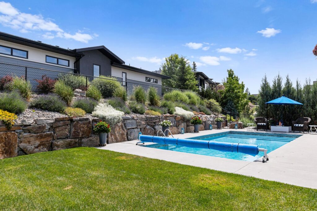 Rectangular pool on a concrete deck below a rock retaining wall planted with grasses, with lawn in the foreground.