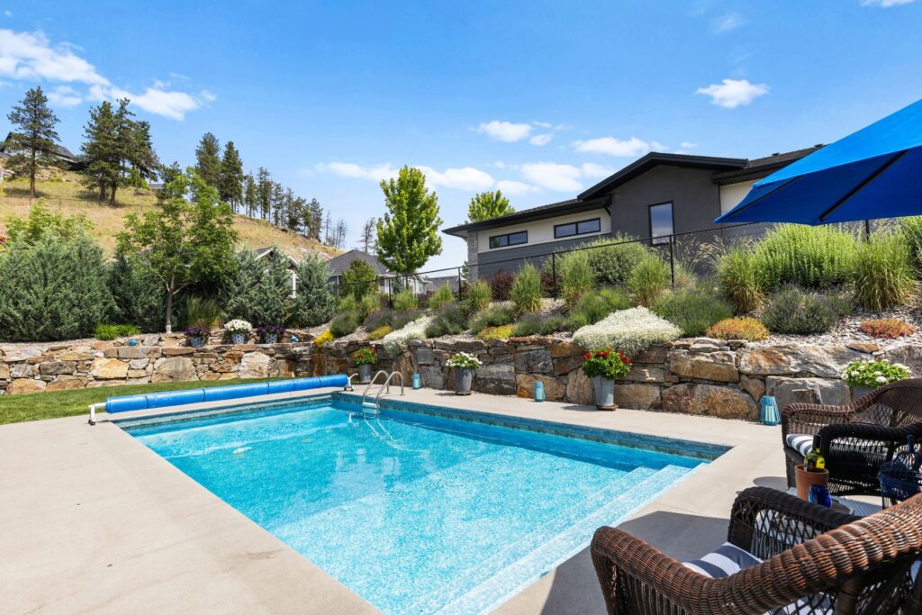 Pool seen from a shaded seating area with wicker chairs and a blue umbrella, houses and dry hillside behind.