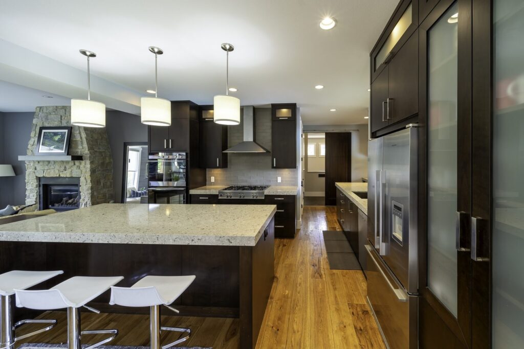 Kitchen with dark cabinetry, wall ovens, a granite island with white stools and a stone fireplace beyond.