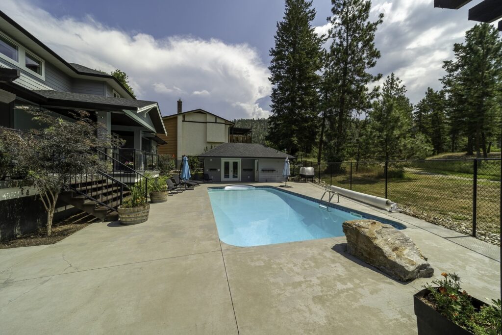 Fenced back garden with an in-ground pool, a concrete surround and mature trees along the boundary.