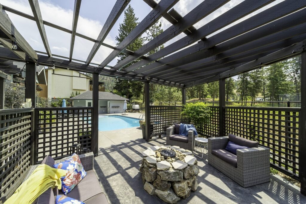 Shaded seating under a lattice pergola beside a back-garden pool, with mature trees along the fence.