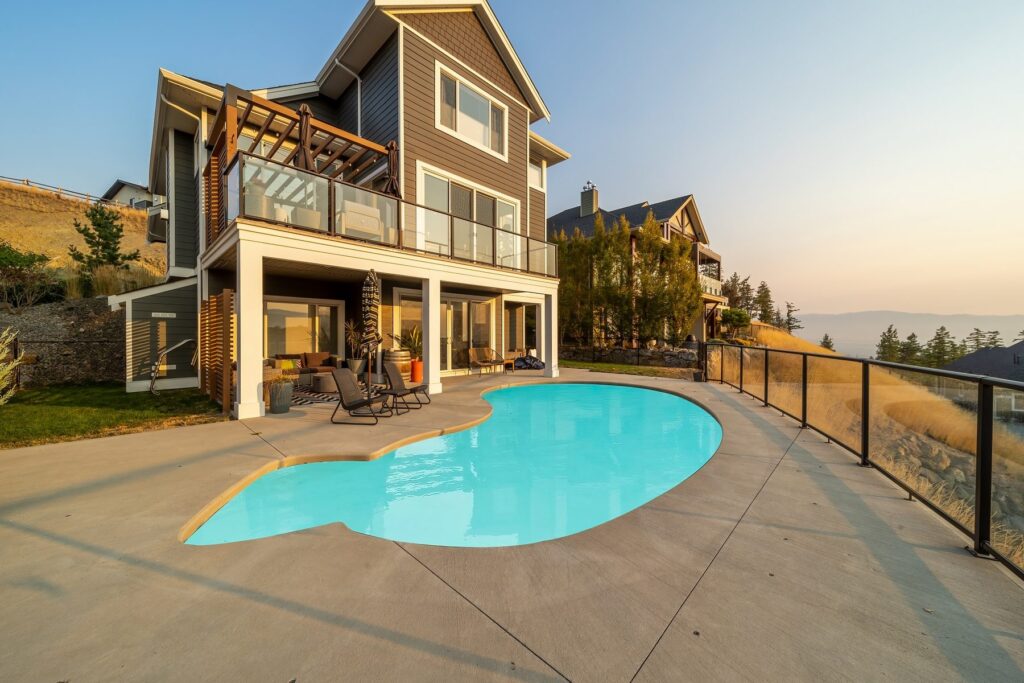 Rear of a house at dusk with its interior lights on, above a lit kidney-shaped pool and concrete deck.