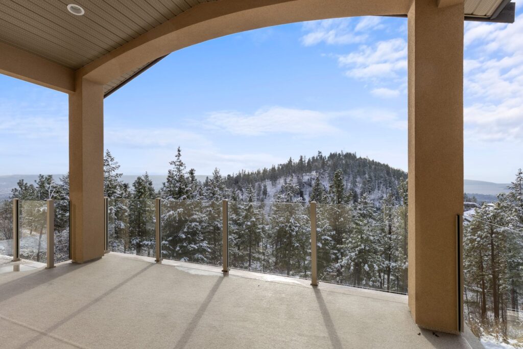 Covered deck with an arched opening and glass railing, looking out over a snow-covered wooded hillside.