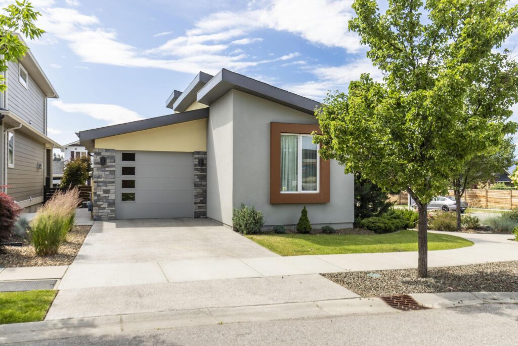Street side of the home in grey and yellow stucco with a skillion roofline, a single-car attached garage with a column of small windows between stacked stone pillars, a concrete driveway and a trimmed lawn.
