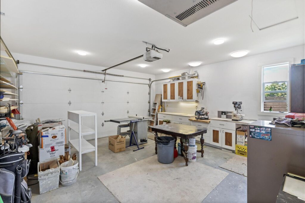 Inside the detached double garage, set up as a workshop: a wide white overhead door with an opener, ceiling heater, white cabinets and counters along the side wall, a table saw, a mitre saw and a drill press.
