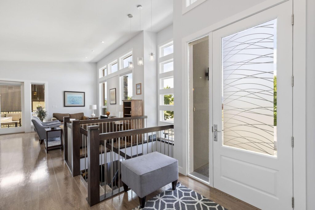 Entry with a white front door with a frosted glass panel and a clear sidelight, a grey ottoman on a geometric rug, and an open stairwell with dark wood and metal-spindle railings leading down to the lower level.