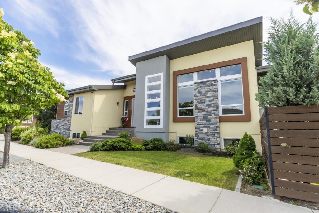 Front of a modern rancher in yellow and grey stucco with a flat overhanging roof, a large brown-framed window wall split by a stacked stone column, a vertical strip of windows, a wood-clad entry with a small Canadian flag, concrete steps, basement windows at grade, evergreen shrubs, a front lawn and a horizontal wood fence.