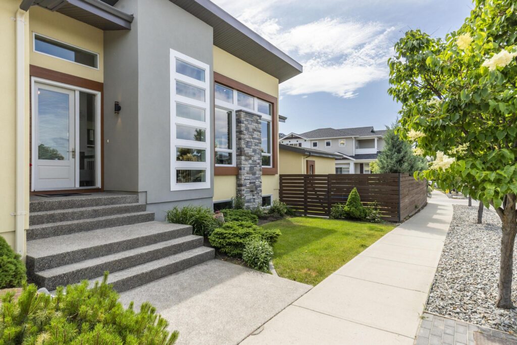 Front entry of a one-storey stucco home with a glass-panel door and transom, wide exposed-aggregate steps, tall stacked windows beside a stone column and basement windows at grade. The detached garage, in matching yellow stucco with brown trim, stands behind the horizontal wood fence.