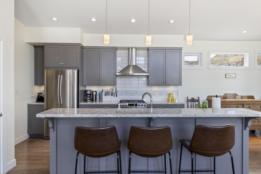 Kitchen with a long quartz island and three brown leather stools, grey shaker cabinets, a French-door stainless fridge, a gas range under a stainless chimney hood, white tile backsplash and three glass pendant lights. Clerestory windows look out to a hillside with houses.