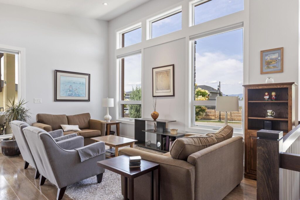 Living room with a high ceiling and tall windows topped by transoms, two grey armchairs, two brown sofas, a wood coffee table and a wood bookcase, with the top of the stair railing in the corner.
