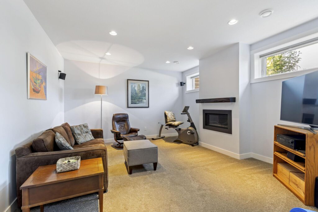 Lower-level family room with a linear gas fireplace under a dark wood mantel, high windows near the ceiling, a sofa, a leather recliner and ottoman, and a television on a wood stand, with carpet and pot lights.