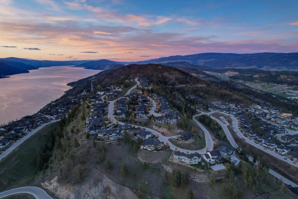 Aerial view of Wilden at dusk: curving streets of hillside homes on the Glenmore Highlands above Okanagan Lake, Kelowna, with the far shore and sunset sky behind